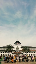 A large group of people is gathered in front of a historic building with unique architectural features, including a central tower. The Indonesian flag is raised above the building, and palm trees are situated on either side. The sky is blue with some clouds.