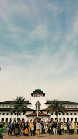 A large group of people is gathered in front of a historic building with unique architectural features, including a central tower. The Indonesian flag is raised above the building, and palm trees are situated on either side. The sky is blue with some clouds.