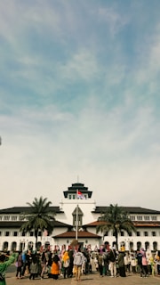 A large group of people is gathered in front of a historic building with unique architectural features, including a central tower. The Indonesian flag is raised above the building, and palm trees are situated on either side. The sky is blue with some clouds.
