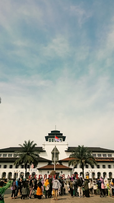 A large group of people is gathered in front of a historic building with unique architectural features, including a central tower. The Indonesian flag is raised above the building, and palm trees are situated on either side. The sky is blue with some clouds.