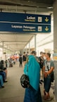 A covered walkway is bustling with people. A blue sign hanging overhead indicates the directions for 'Ticket Counter' and 'Customer Service' in English and another language, with arrows pointing downwards. Many individuals are visible in the walkway, some sitting, some standing, and some wearing face masks. A woman in a turquoise headscarf and black backpack stands in the foreground.
