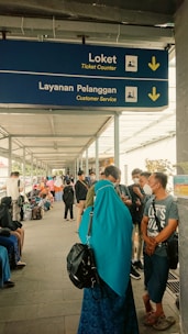 A covered walkway is bustling with people. A blue sign hanging overhead indicates the directions for 'Ticket Counter' and 'Customer Service' in English and another language, with arrows pointing downwards. Many individuals are visible in the walkway, some sitting, some standing, and some wearing face masks. A woman in a turquoise headscarf and black backpack stands in the foreground.