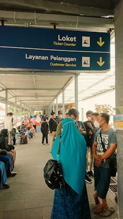A covered walkway is bustling with people. A blue sign hanging overhead indicates the directions for 'Ticket Counter' and 'Customer Service' in English and another language, with arrows pointing downwards. Many individuals are visible in the walkway, some sitting, some standing, and some wearing face masks. A woman in a turquoise headscarf and black backpack stands in the foreground.