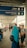 A covered walkway is bustling with people. A blue sign hanging overhead indicates the directions for 'Ticket Counter' and 'Customer Service' in English and another language, with arrows pointing downwards. Many individuals are visible in the walkway, some sitting, some standing, and some wearing face masks. A woman in a turquoise headscarf and black backpack stands in the foreground.