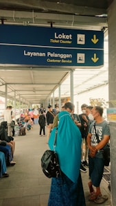 A covered walkway is bustling with people. A blue sign hanging overhead indicates the directions for 'Ticket Counter' and 'Customer Service' in English and another language, with arrows pointing downwards. Many individuals are visible in the walkway, some sitting, some standing, and some wearing face masks. A woman in a turquoise headscarf and black backpack stands in the foreground.
