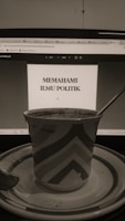 A sepia-toned photo displays a computer screen showing a document titled 'Memahami Ilmu Politik'. In front of the screen, there is a patterned mug placed on a saucer with a spoon beside it.