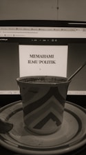A sepia-toned photo displays a computer screen showing a document titled 'Memahami Ilmu Politik'. In front of the screen, there is a patterned mug placed on a saucer with a spoon beside it.