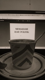 A sepia-toned photo displays a computer screen showing a document titled 'Memahami Ilmu Politik'. In front of the screen, there is a patterned mug placed on a saucer with a spoon beside it.