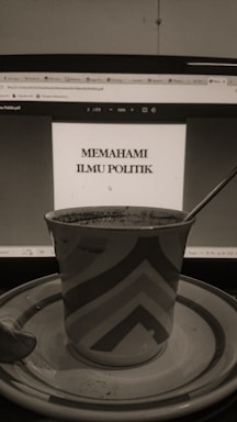 A sepia-toned photo displays a computer screen showing a document titled 'Memahami Ilmu Politik'. In front of the screen, there is a patterned mug placed on a saucer with a spoon beside it.