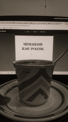 A sepia-toned photo displays a computer screen showing a document titled 'Memahami Ilmu Politik'. In front of the screen, there is a patterned mug placed on a saucer with a spoon beside it.