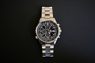 A stainless steel wristwatch with a complex dial featuring multiple sub-dials and a date function, set against a black background. The watch has a polished metal strap and a well-illuminated face highlighting intricate details.