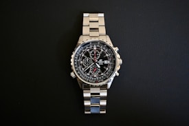 A stainless steel wristwatch with a complex dial featuring multiple sub-dials and a date function, set against a black background. The watch has a polished metal strap and a well-illuminated face highlighting intricate details.