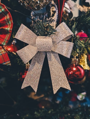 A glittery silver bow is prominently displayed on a Christmas tree, surrounded by festive ornaments including shiny red baubles and a plaid-patterned bow. The branches of the tree are dark green, adding a contrasting backdrop to the bright decorations.
