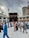 A large gathering of people, many wearing traditional Islamic clothing, surrounds the Kaaba in the courtyard of the Masjid al-Haram. The sky is overcast, and there are tall buildings and cranes visible in the background.