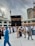 A large gathering of people, many wearing traditional Islamic clothing, surrounds the Kaaba in the courtyard of the Masjid al-Haram. The sky is overcast, and there are tall buildings and cranes visible in the background.