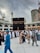 A large gathering of people, many wearing traditional Islamic clothing, surrounds the Kaaba in the courtyard of the Masjid al-Haram. The sky is overcast, and there are tall buildings and cranes visible in the background.