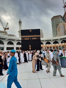 A large gathering of people, many wearing traditional Islamic clothing, surrounds the Kaaba in the courtyard of the Masjid al-Haram. The sky is overcast, and there are tall buildings and cranes visible in the background.