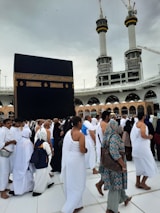 A crowd of people performing the Tawaf, a religious ritual, around the Kaaba in Mecca. The atmosphere is busy and reverent, with men and women dressed predominantly in white robes.
