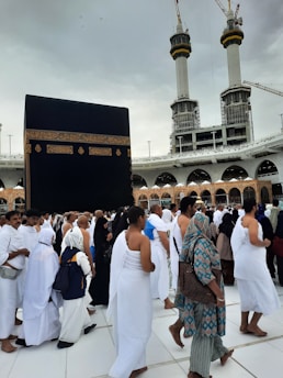 A crowd of people performing the Tawaf, a religious ritual, around the Kaaba in Mecca. The atmosphere is busy and reverent, with men and women dressed predominantly in white robes.