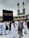 A crowd of people performing the Tawaf, a religious ritual, around the Kaaba in Mecca. The atmosphere is busy and reverent, with men and women dressed predominantly in white robes.