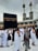 A crowd of people performing the Tawaf, a religious ritual, around the Kaaba in Mecca. The atmosphere is busy and reverent, with men and women dressed predominantly in white robes.