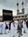 A crowd of people performing the Tawaf, a religious ritual, around the Kaaba in Mecca. The atmosphere is busy and reverent, with men and women dressed predominantly in white robes.