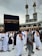 A crowd of people performing the Tawaf, a religious ritual, around the Kaaba in Mecca. The atmosphere is busy and reverent, with men and women dressed predominantly in white robes.