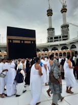 A crowd of people performing the Tawaf, a religious ritual, around the Kaaba in Mecca. The atmosphere is busy and reverent, with men and women dressed predominantly in white robes.