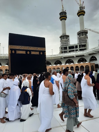 A crowd of people performing the Tawaf, a religious ritual, around the Kaaba in Mecca. The atmosphere is busy and reverent, with men and women dressed predominantly in white robes.