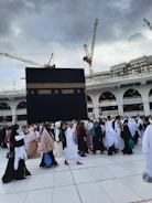 A large gathering of people in white and various colored garments are walking on tiled ground. In the background, the Kaaba, a large black cube with gold embroidery, dominates the scene. Construction cranes and a multi-story building with arches are visible under a cloudy sky.