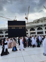 A large gathering of people in white and various colored garments are walking on tiled ground. In the background, the Kaaba, a large black cube with gold embroidery, dominates the scene. Construction cranes and a multi-story building with arches are visible under a cloudy sky.