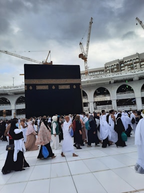 A large gathering of people in white and various colored garments are walking on tiled ground. In the background, the Kaaba, a large black cube with gold embroidery, dominates the scene. Construction cranes and a multi-story building with arches are visible under a cloudy sky.