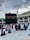 A large gathering of people in white and various colored garments are walking on tiled ground. In the background, the Kaaba, a large black cube with gold embroidery, dominates the scene. Construction cranes and a multi-story building with arches are visible under a cloudy sky.
