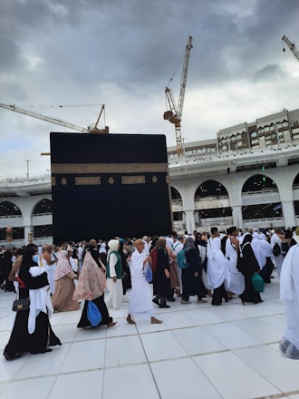 A large gathering of people in white and various colored garments are walking on tiled ground. In the background, the Kaaba, a large black cube with gold embroidery, dominates the scene. Construction cranes and a multi-story building with arches are visible under a cloudy sky.
