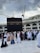 A large gathering of people in white and various colored garments are walking on tiled ground. In the background, the Kaaba, a large black cube with gold embroidery, dominates the scene. Construction cranes and a multi-story building with arches are visible under a cloudy sky.