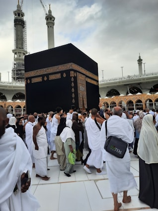 A large gathering of people dressed in traditional white garments surrounding the Kaaba, a significant religious structure, with a partially cloudy sky and tall minarets in the background.