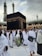 A large gathering of people dressed in traditional white garments surrounding the Kaaba, a significant religious structure, with a partially cloudy sky and tall minarets in the background.