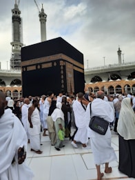 A large gathering of people dressed in traditional white garments surrounding the Kaaba, a significant religious structure, with a partially cloudy sky and tall minarets in the background.