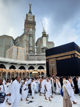 A large crowd of people dressed in white garments walk around a prominent black structure with gold details, set against the backdrop of a towering clock tower and surrounding buildings. The scene appears to be a religious gathering in an expansive courtyard.