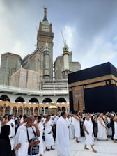 A large group of people, many dressed in white robes, are gathered around the Kaaba, a black cube structure adorned with golden details. The towering Abraj Al Bait Clock Tower is visible in the background, with its large clock and decorative spire. The scene conveys a sense of a spiritual gathering.