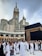 A large group of people, many dressed in white robes, are gathered around the Kaaba, a black cube structure adorned with golden details. The towering Abraj Al Bait Clock Tower is visible in the background, with its large clock and decorative spire. The scene conveys a sense of a spiritual gathering.