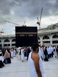 A large crowd of people wearing traditional white garments gather around the Kaaba, a cuboid structure in the courtyard of the Great Mosque of Mecca. Overhead, several towering cranes are visible against a cloudy sky.