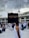 A large crowd of people wearing traditional white garments gather around the Kaaba, a cuboid structure in the courtyard of the Great Mosque of Mecca. Overhead, several towering cranes are visible against a cloudy sky.