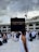 A large crowd of people wearing traditional white garments gather around the Kaaba, a cuboid structure in the courtyard of the Great Mosque of Mecca. Overhead, several towering cranes are visible against a cloudy sky.