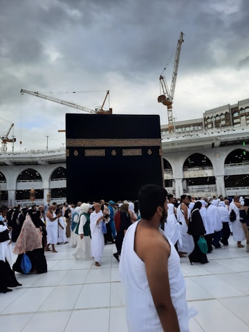 A large crowd of people wearing traditional white garments gather around the Kaaba, a cuboid structure in the courtyard of the Great Mosque of Mecca. Overhead, several towering cranes are visible against a cloudy sky.