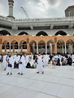 A large group of people dressed in white garments gather in an open area with a tiled floor. The background features an ornate building with arched architecture and a tower under gray, cloudy skies. People appear to be engaged in activities such as walking, talking, or sitting.