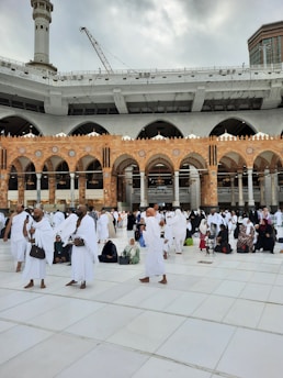 A large group of people dressed in white garments gather in an open area with a tiled floor. The background features an ornate building with arched architecture and a tower under gray, cloudy skies. People appear to be engaged in activities such as walking, talking, or sitting.