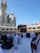 A large crowd of people, many dressed in white robes, gather around the Kaaba in an open courtyard. The surrounding architecture includes arched structures and a towering building with a large clock. The sky is clear and blue.
