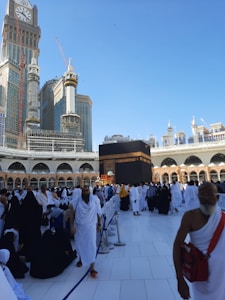 A large crowd of people, many dressed in white robes, gather around the Kaaba in an open courtyard. The surrounding architecture includes arched structures and a towering building with a large clock. The sky is clear and blue.
