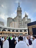 A large crowd of people dressed in traditional attire gather around a significant religious site with a tall clock tower and impressive architectural structures in the background. The scene is bustling with activity and a sense of reverence.